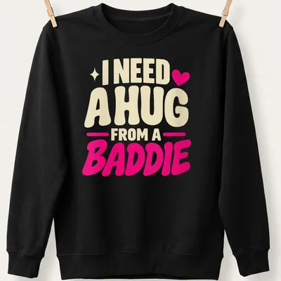 Bold playful text saying 'I Need A Hug From A Baddie' in cream, pink, and red — printed on a unisex sweatshirt