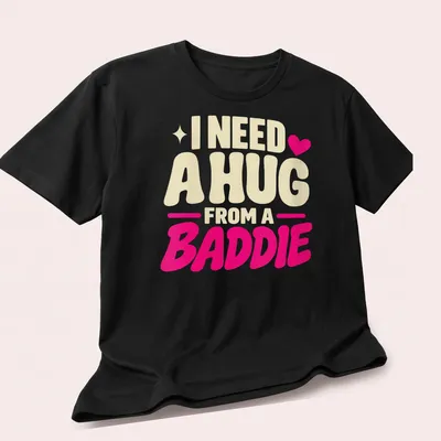 Bold playful text saying 'I Need A Hug From A Baddie' in cream, pink, and red — printed on a unisex t-shirt (angled view)