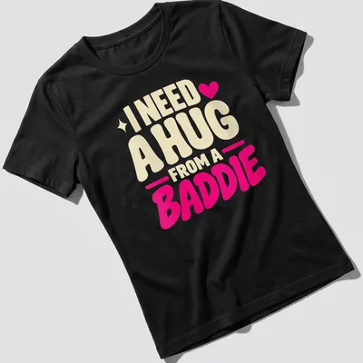 Bold playful text saying 'I Need A Hug From A Baddie' in cream, pink, and red — printed on a women's shirt
