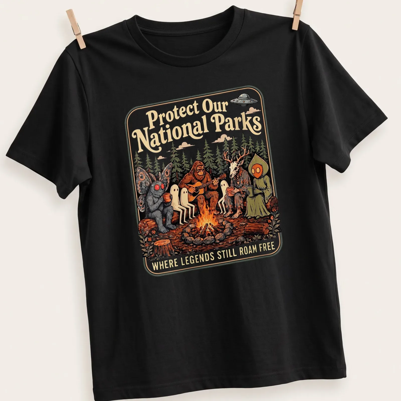 Vintage illustration of Bigfoot and Mothman around a campfire with text about protecting national parks. — printed on a unisex t-shirt